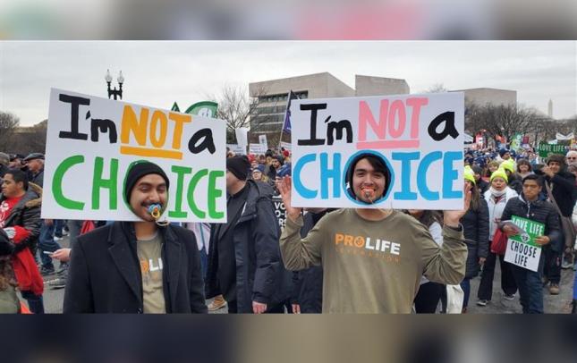 Two young men at a march holding large hand-lettered signs reading “I’m not a choice.”